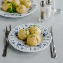 Trzy pyzy ziemniaczane na talerzu z niebieskim wzorem, udekorowane natką pietruszki. Obok widelec i nóż.