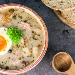 Tradycyjny żurek w butelce, podany z jajkiem i natką pietruszki. Obok kromki chleba i drewniana łyżka.