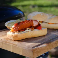 Pyszna kiełbasa na grilla w bułce z warzywami, przygotowana na drewnianej desce obok grilla.
