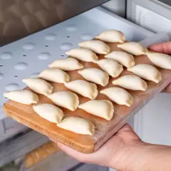 Ręce wkładają deskę z surowymi pierogami do zamrażarki. Gotowe do mrożenia pierogi czekają na swój czas.