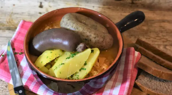 Krupniok i kaszanka w glinianym naczyniu z ziemniakami i kapustą, obok kromki chleba.