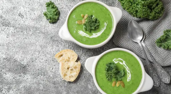 Dwie miseczki zielonego kremu z jarmużu, ozdobione listkiem jarmużu i pestkami dyni. Czy można zabielić zupę jogurtem naturalnym? Obok pieczywo i łyżeczki.
