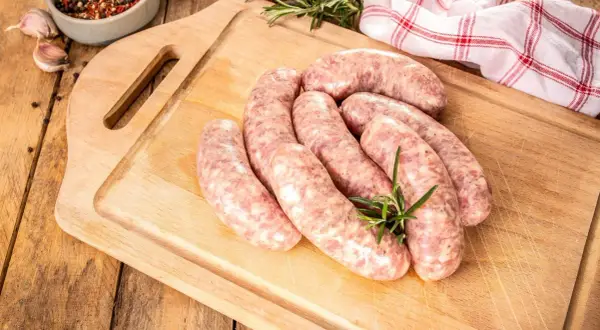 Kilka surowych kiełbasek białych parzonych ułożonych na desce, ozdobionych gałązką rozmarynu. Obok miseczka z przyprawami i czosnek.