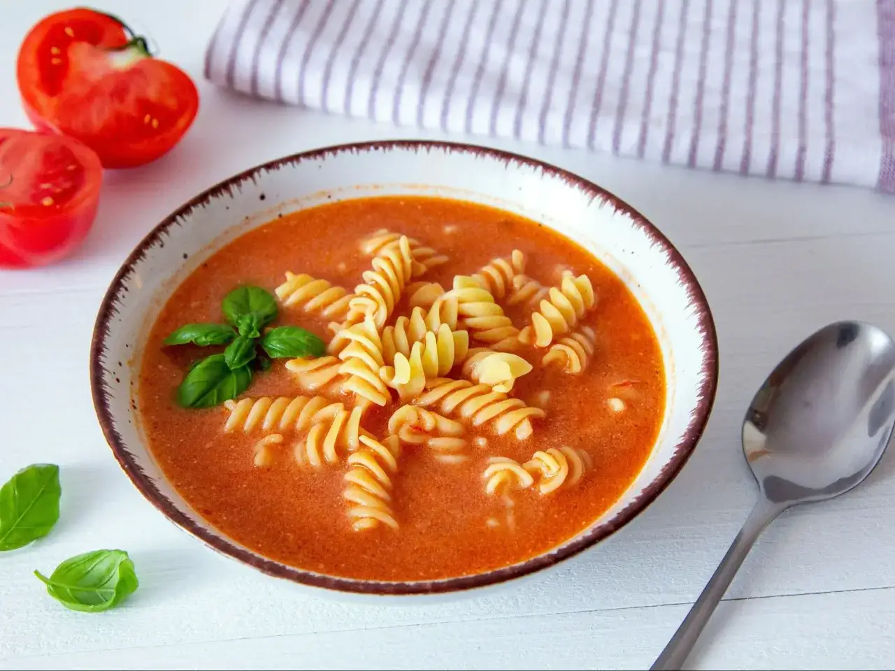 Pomidorowa z rosołu z makaronem fusilli i bazylią. Obok miski świeże pomidory i łyżka.