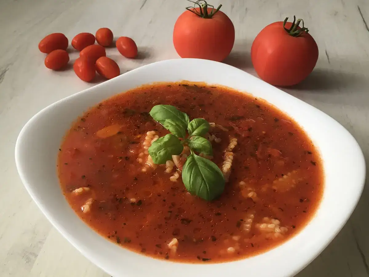 Aksamitna zupa pomidorowa na kostce, udekorowana bazylią, z makaronem. Obok leżą świeże pomidory.