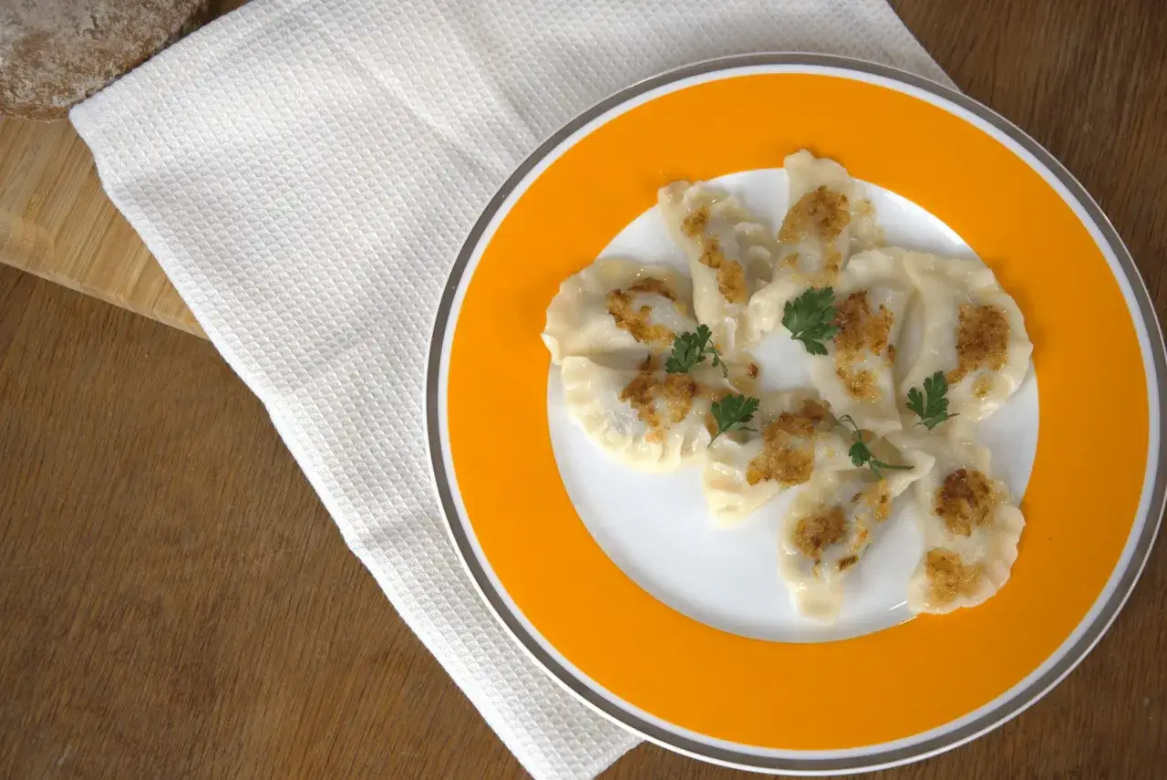 Pyszne pierogi z gęsiną, posypane skwarkami i udekorowane natką pietruszki, na pomarańczowym talerzu.