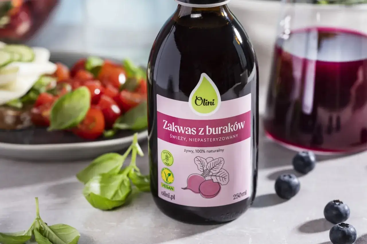 Butelka naturalnego zakwasu z buraków Olini, obok talerza z pomidorami i bazylią. Zdrowy, kiszony barszcz czerwony w płynie.